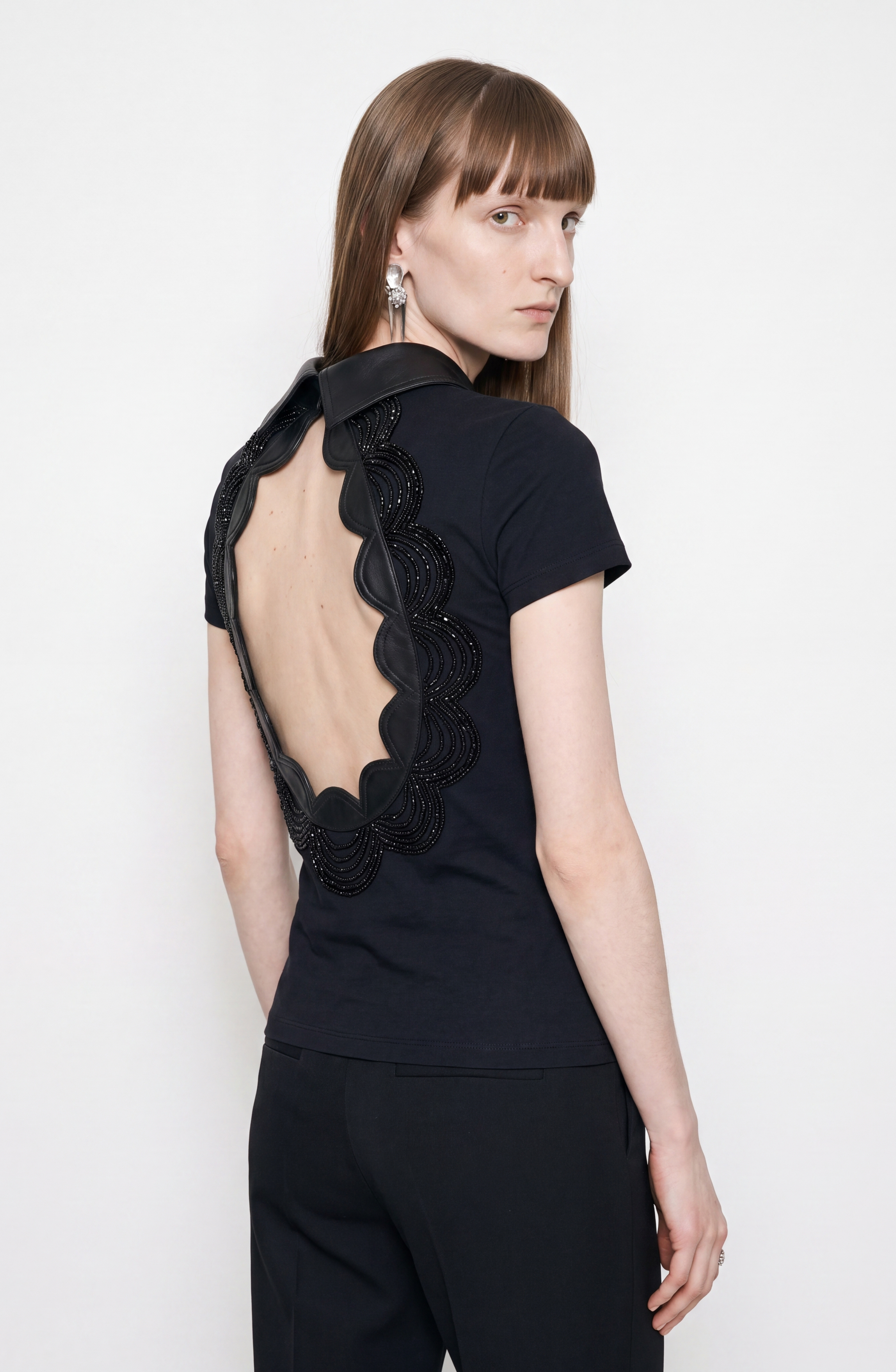 A woman with straight brown hair wears the LOULOU Scalloped Open Back Top, a black cotton-blend fitted piece featuring a large, scalloped open back and a sleek leather collar detail. Paired with black pants, she stands against a plain white background, looking over her shoulder and showcasing dangling silver earrings that enhance her modern style.