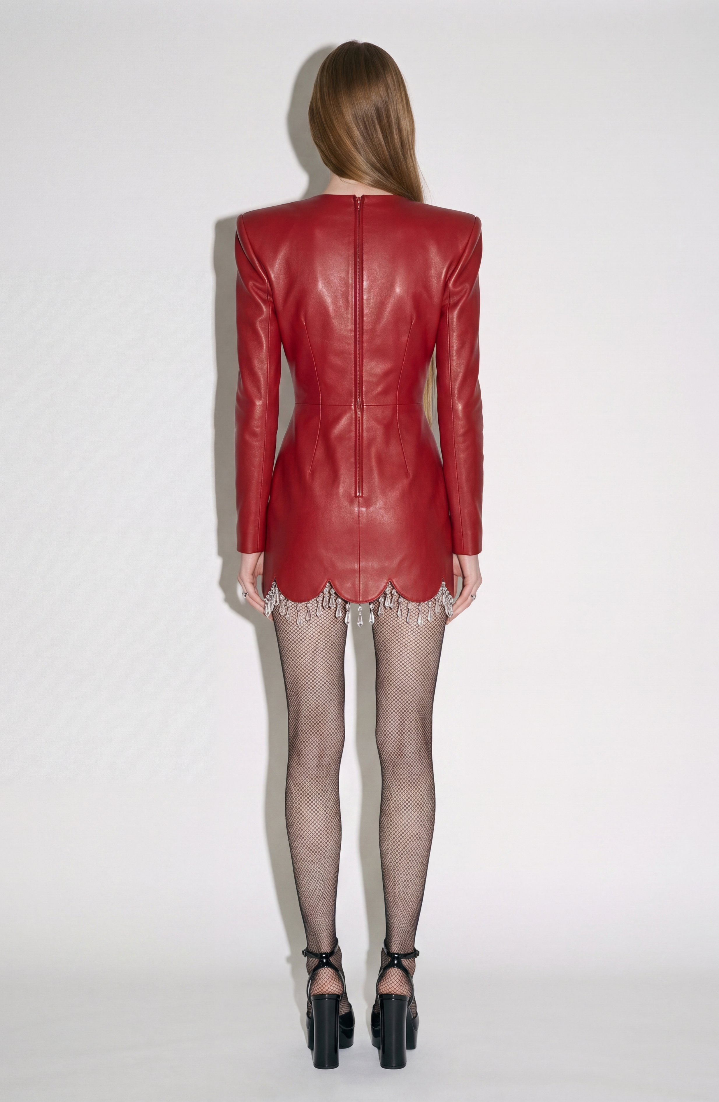 A woman faces away in the LOULOU Scarlet Leather Dress—a fitted red lamb leather mini with sculptural shoulders and silver hem embellishments—styled with fishnet tights and black heels, set against a plain white background.