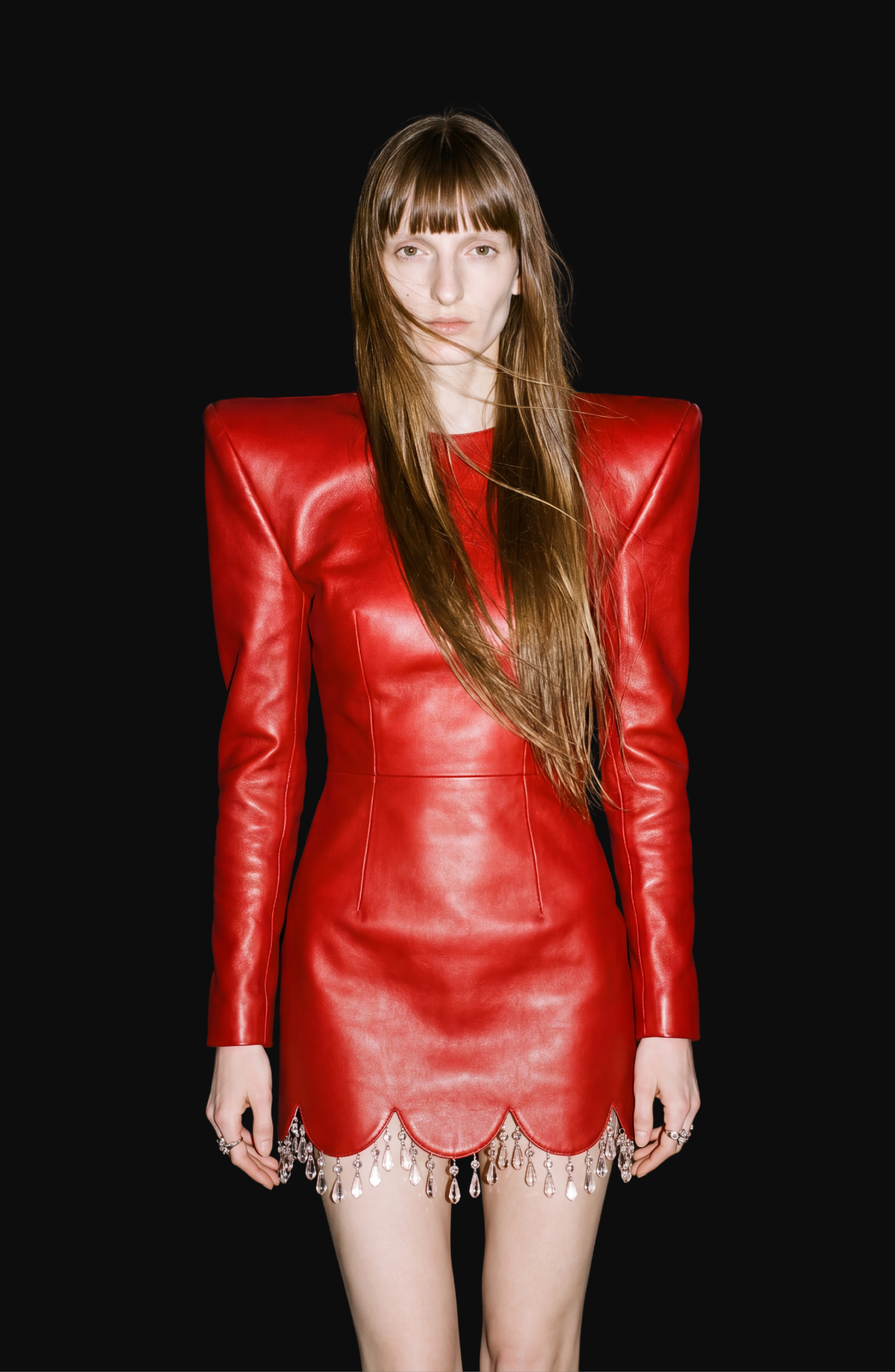 A woman with long brown hair stands against a black background, wearing the LOULOU Scarlet Leather Dress—a lamb leather mini dress with sculptural shoulders and a scalloped hem adorned with sparkling crystal details.