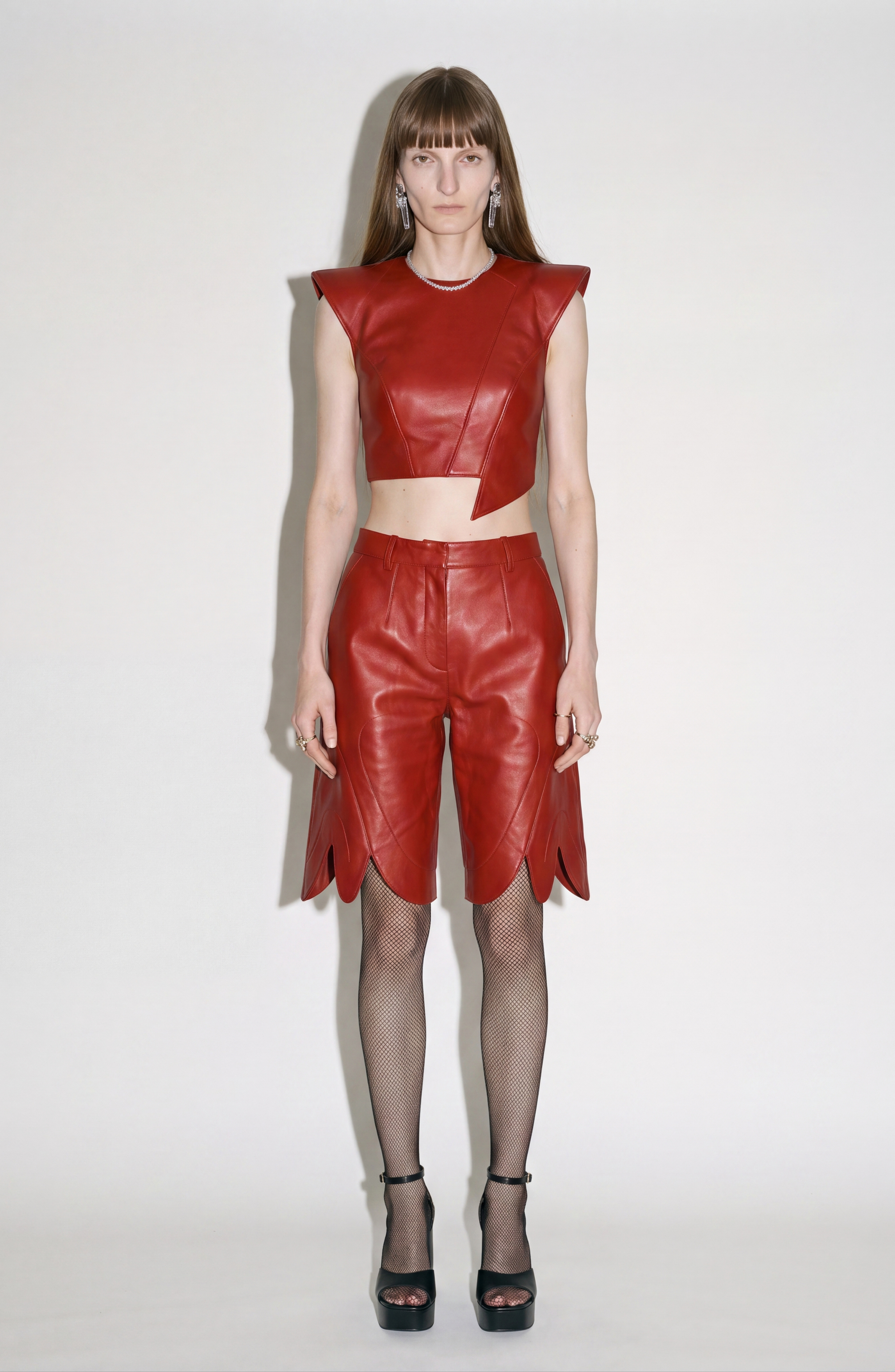 A person poses against a white background in the LOULOU Red Leather Top paired with matching red leather shorts, styled with black fishnet stockings and black high-heeled sandals.