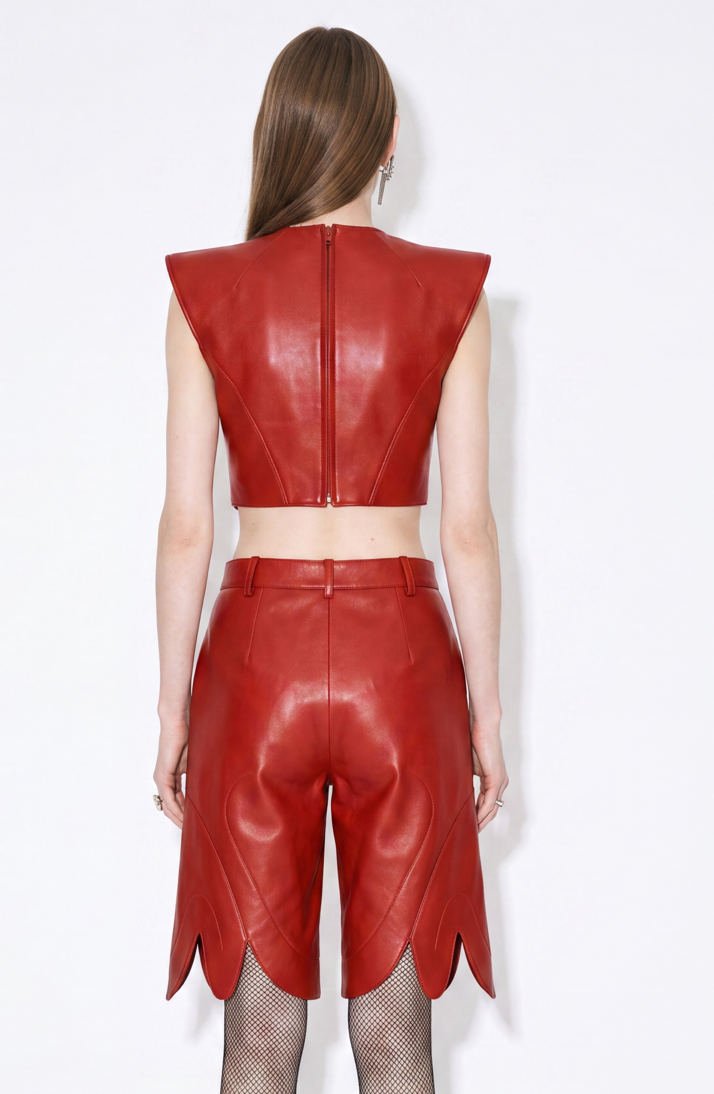 A person with long brown hair is seen from the back wearing the LOULOU Red Leather Top, a sleeveless lamb leather piece with sculptural cap shoulders, paired with matching high-waisted scallop-hem shorts against a white background.