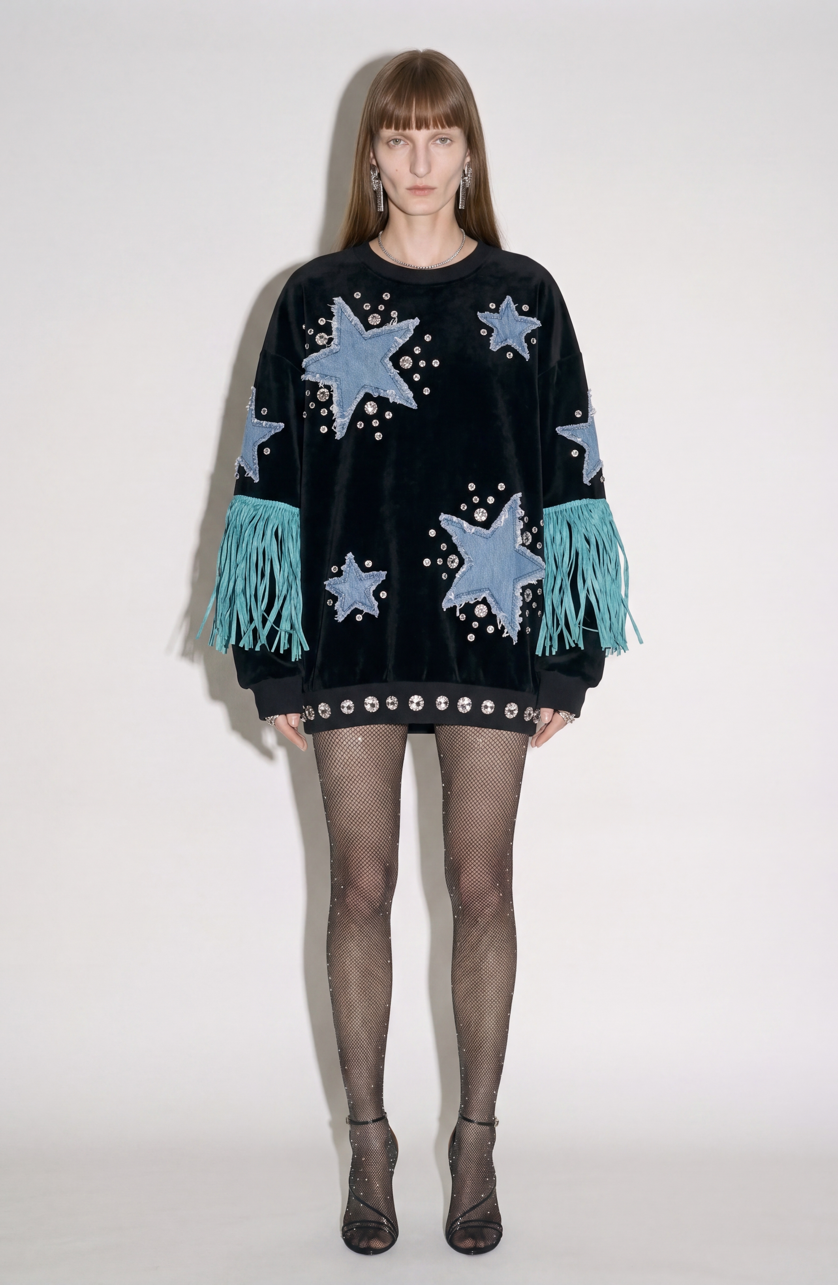 A person is wearing the LOULOU Star Appliqué Velvet Sweatshirt—an oversized black velvet top adorned with denim star appliqués, silver studs, and teal fringe on the sleeves—paired with sheer polka-dot tights and black heels, standing against a plain white background.