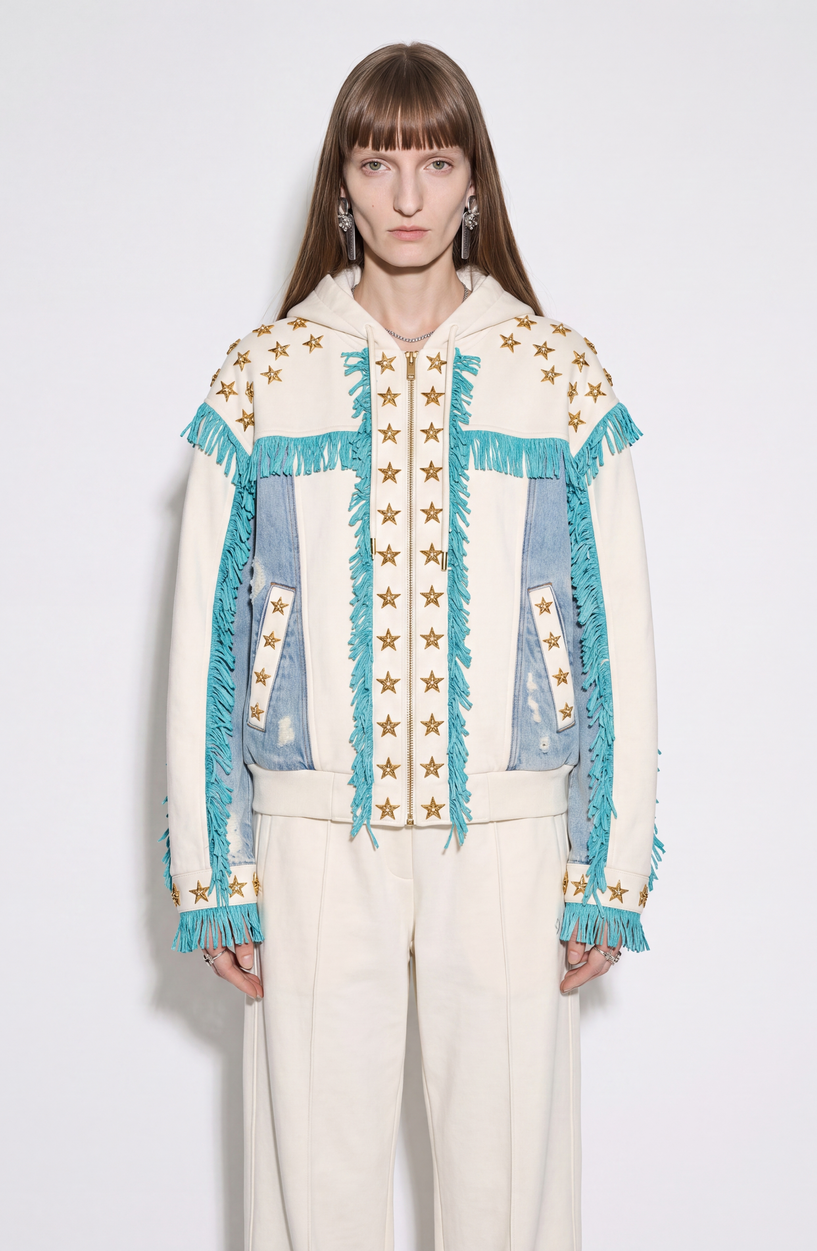 A person wearing the LOULOU Star Studded Fringed Zip Hoodie—featuring cream and denim panels, turquoise fringe, gold star details, and distressed accents—stands against a plain white background, looking directly at the camera. The cohesive look is completed with matching cream pants.