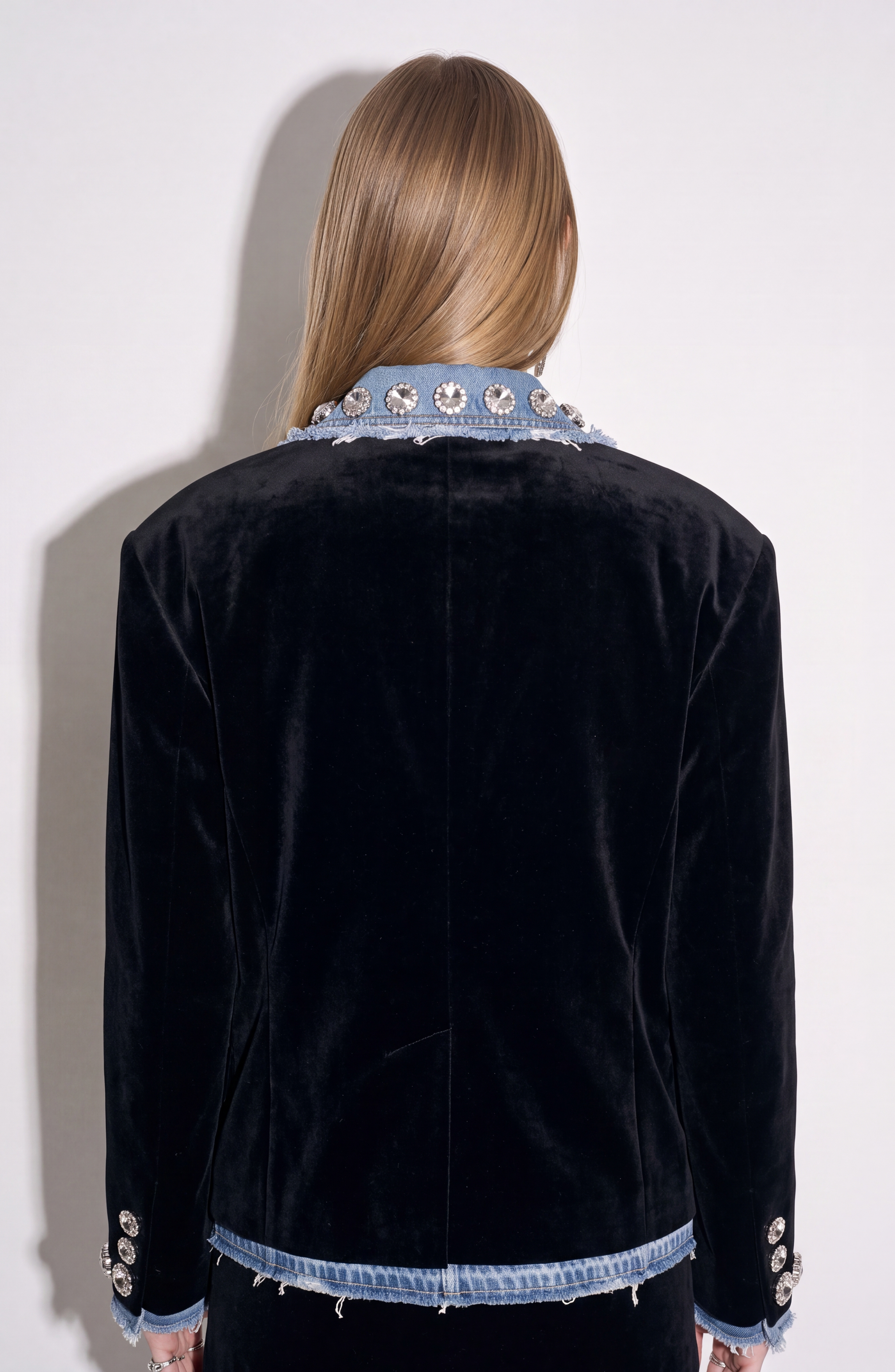 A person with long, straight hair, seen from behind, is wearing the LOULOU Velvet And Denim Crystal Blazer featuring decorative buttons and rhinestone embellishments on the collar and cuffs.