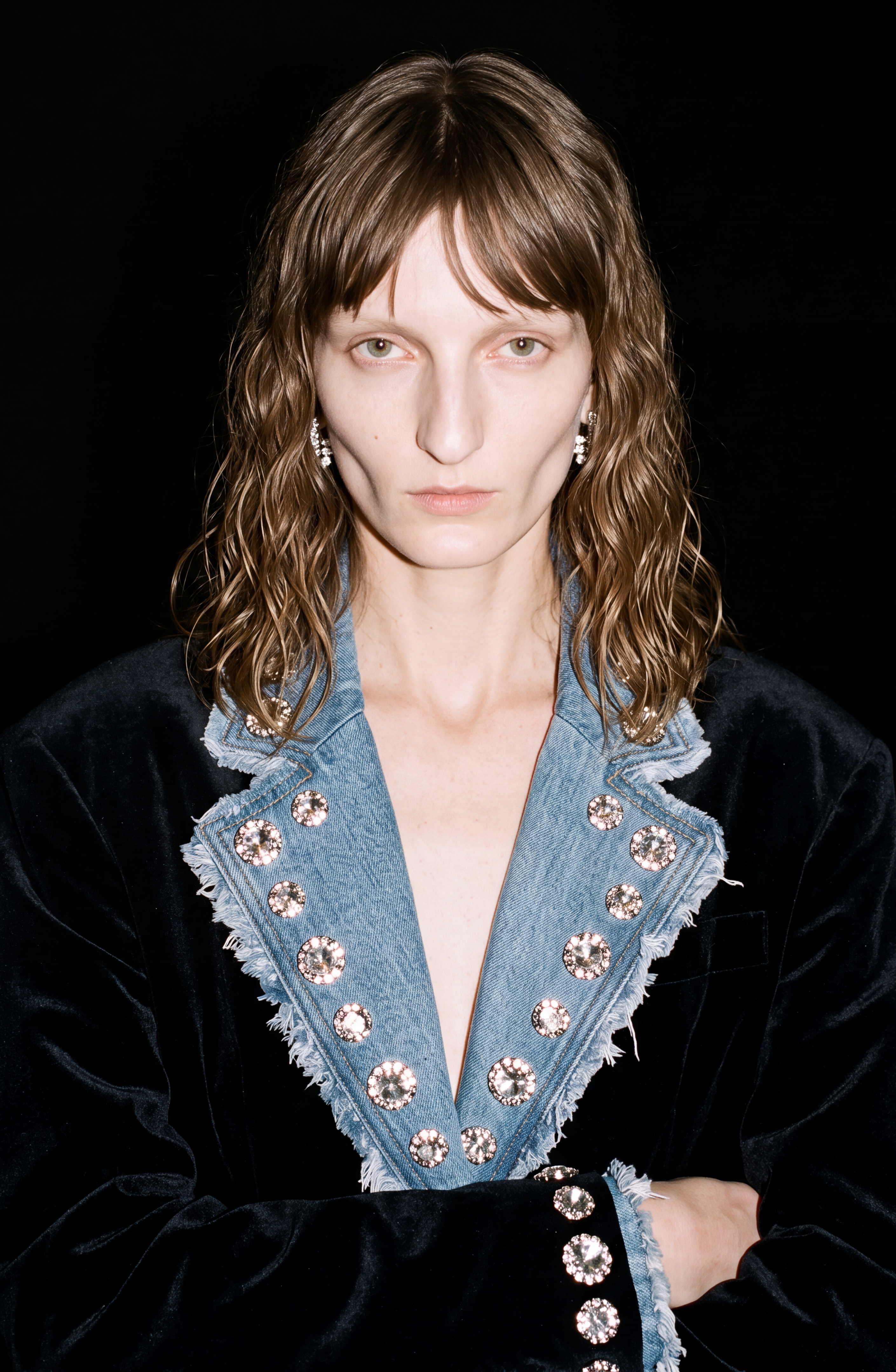 A person with wavy brown hair stands against a black background wearing the LOULOU Velvet And Denim Crystal Blazer, a dark velvet piece highlighted by a bold blue denim lapel embellished with sparkling glass crystals, their expression serious.