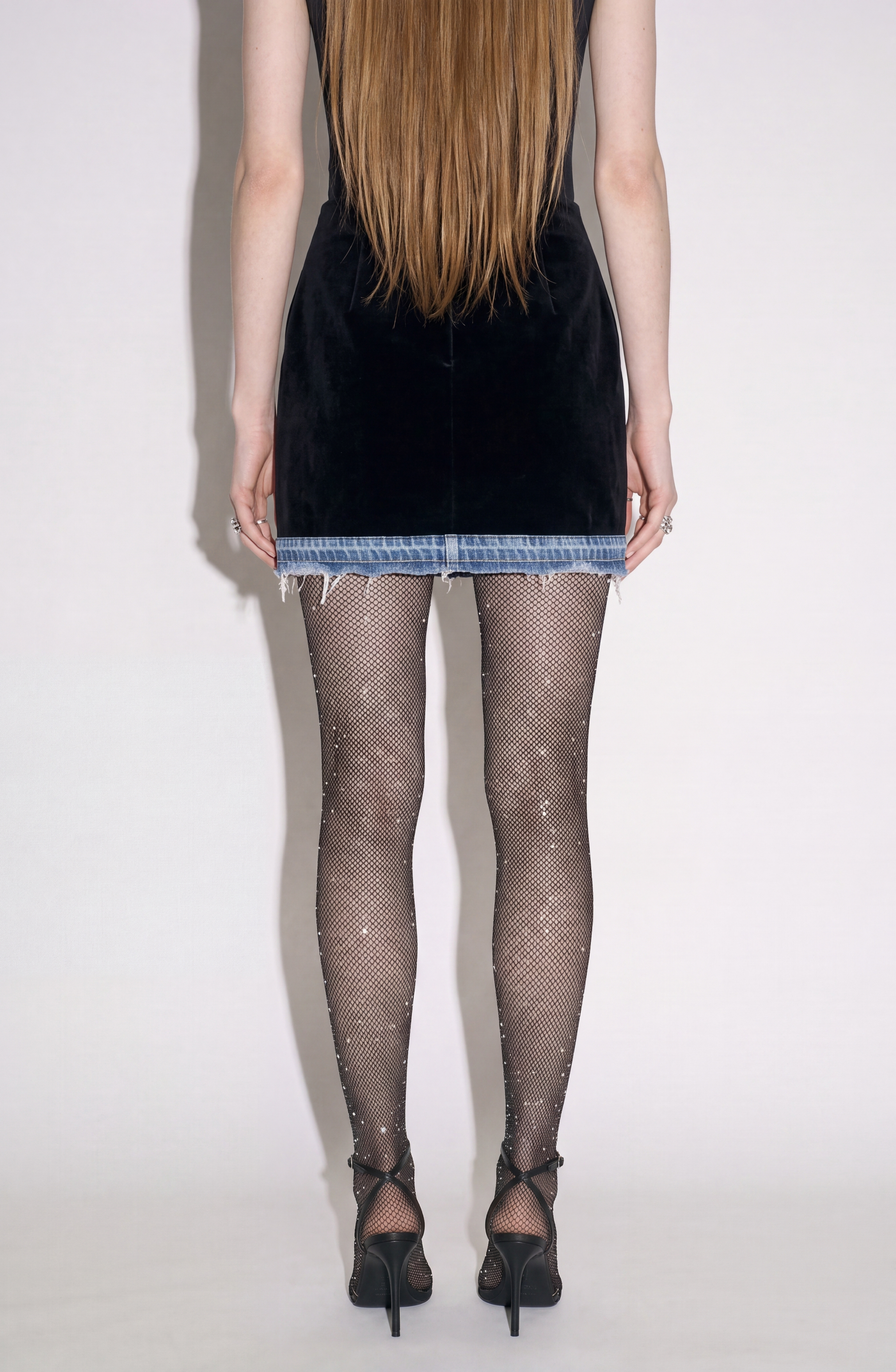 A person stands facing away, wearing the LOULOU Velvet And Denim Mini Skirt paired with sparkly fishnet tights and black high heels. Their long, straight hair falls down their back.