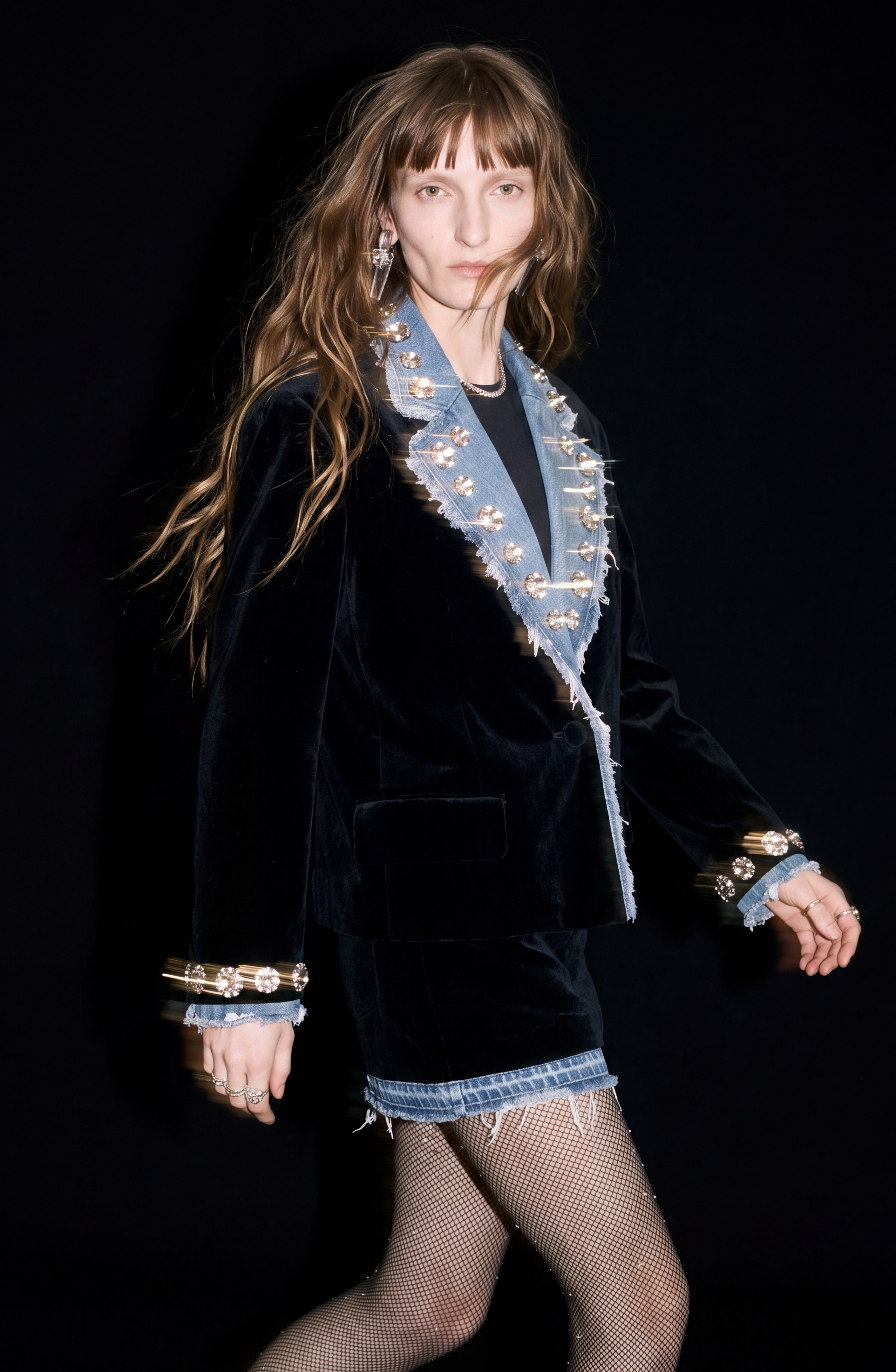 A long-haired model with bangs showcases the LOULOU Velvet And Denim Mini Skirt paired with a coordinating black velvet jacket, both accented by distressed denim lapels and cuffs encrusted with sparkling rhinestones. She completes her look with fishnet stockings and statement earrings, set against a black backdrop.