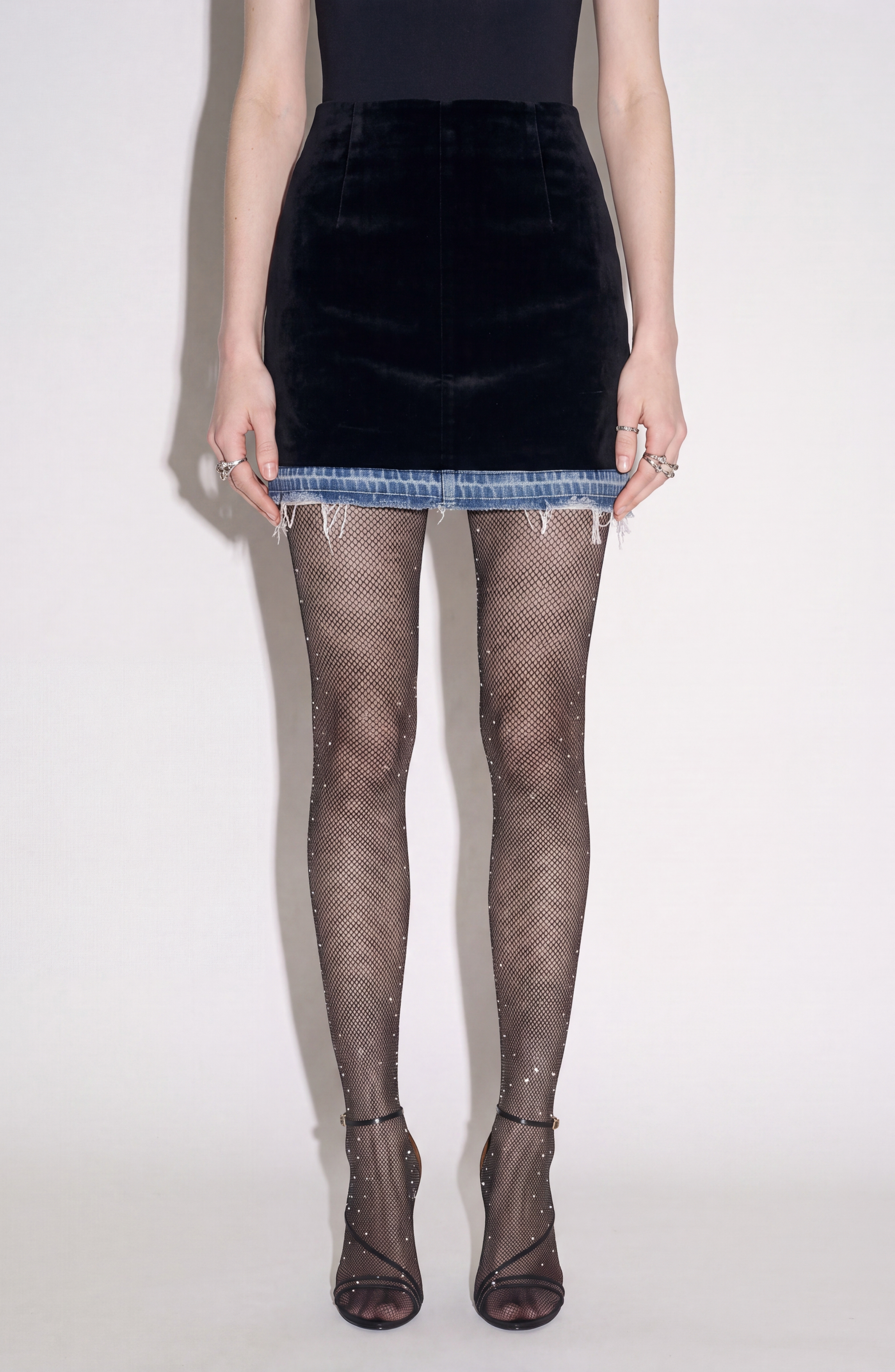 A person stands against a plain light background, wearing a black sleeveless top, sheer black fishnet tights embellished with rhinestones, open-toe heels, and several rings. The edgy look features the LOULOU Velvet And Denim Mini Skirt—a striking piece that combines a frayed denim hem with soft velvet accents for bold textural contrast.